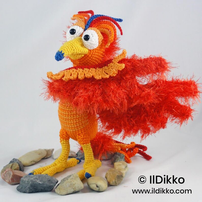 May include: A crocheted phoenix bird toy with a bright orange body, yellow legs, and long, shaggy red wings. It features large white eyes with black pupils, a yellow beak with a blue tip, and blue and red crest feathers. Text reads: &copy; IlDikko www.ildikko.com