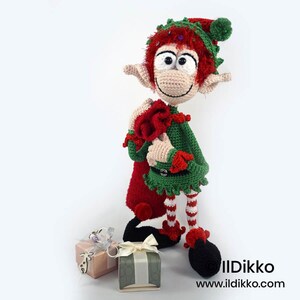 May include: A crocheted elf doll with red hair, a green tunic, and red and white striped leggings, carrying a red sack over its shoulder. The elf wears a green hat with a pom-pom and stands next to two small gift boxes. Text reads Ildikko www.ildikko.com.