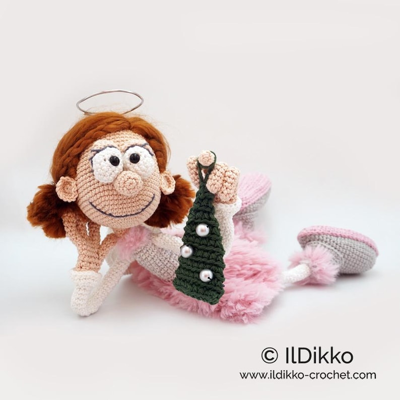 May include: A crocheted doll with auburn hair, a silver wire halo, and a pink fuzzy skirt. The doll has large white eyes, a round nose, and is holding a small dark green triangular ornament decorated with three white beads. Text reads ILDikko.