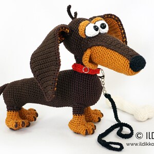 May include: A crocheted dachshund dog with brown and tan yarn. The dog has large, expressive eyes, a red collar, and a black leash. A white bone is next to the dog. The dog is handmade and has a whimsical appearance.
