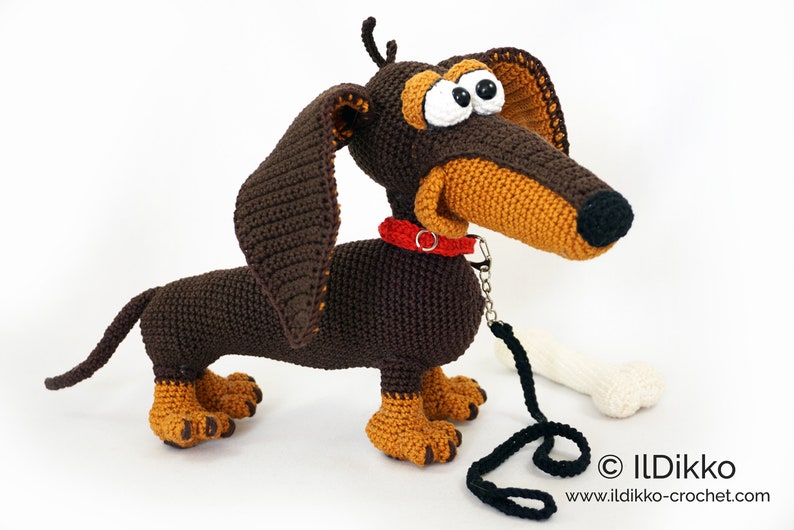May include: A crocheted dachshund dog with brown and tan yarn. The dog has large, expressive eyes, a red collar, and a black leash. A white bone is next to the dog. The dog is handmade and has a whimsical appearance.