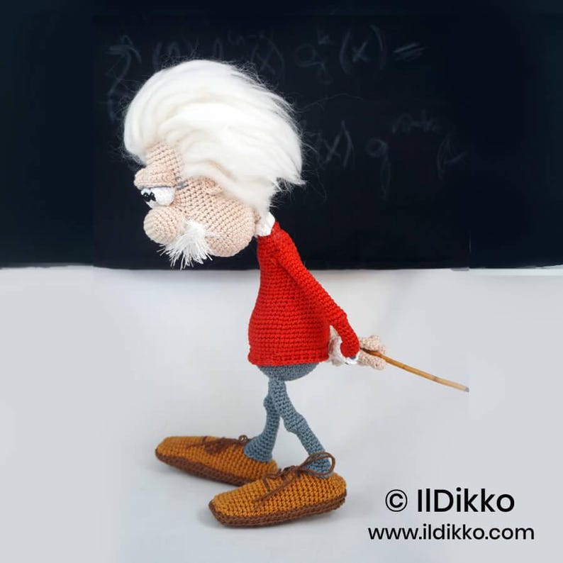 May include: A crocheted doll of an elderly person with wild white hair, a mustache, and glasses. The figure wears a red long-sleeved shirt and grey trousers, with brown shoes. The doll holds a thin wooden pointer behind its back. Text reads: &copy; IlDikko www.ildikko.com
