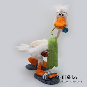 May include: A white crocheted goose wearing green and orange sandals, sunglasses, and a green towel. The goose has a long neck and a blue flower in its beak. The goose is standing on a white background.