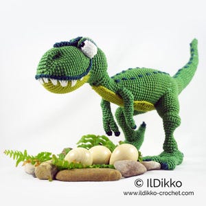 Może przedstawiać: Zielona pluszowa zabawka dinozaura z szerokim uśmiechem i dużymi oczami. Dinozaur stoi na łóżku z brązowych kamieni i zielonych paproci z czterema białymi jajami.
