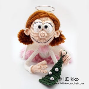 May include: A crocheted doll with auburn hair, a silver wire halo, and a pink skirt. The doll has large white eyes, a stitched smile, and holds a dark green crocheted Christmas tree ornament with three silver beads. Text reads: ILDikko ildikko-crochet.com
