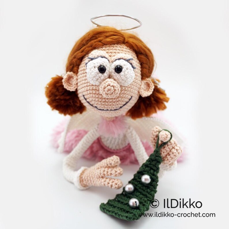 May include: A crocheted doll with auburn hair, a silver wire halo, and a pink skirt. The doll has large white eyes, a stitched smile, and holds a dark green crocheted Christmas tree ornament with three silver beads. Text reads: ILDikko ildikko-crochet.com