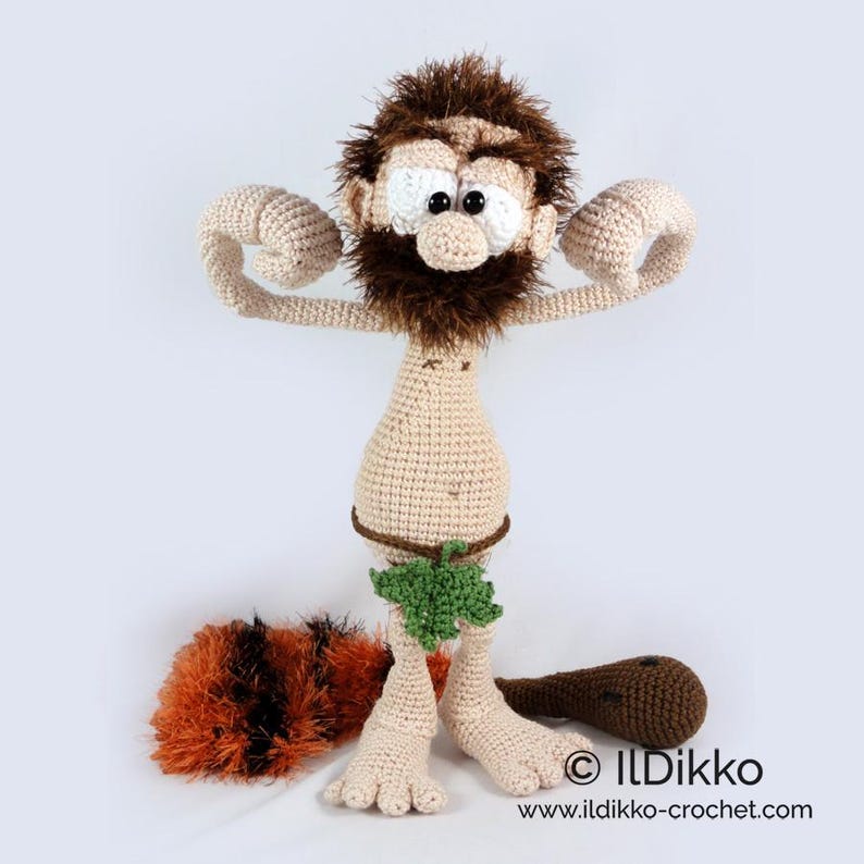 May include: A crocheted caveman doll with beige skin, a dark brown fuzzy beard and hair, and large white eyes with black pupils. The figure wears a green leaf-shaped loincloth and holds a brown club, with a separate orange and black fuzzy club.