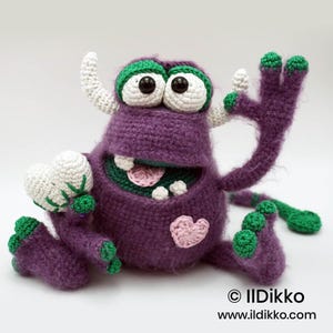Puede incluir: Un juguete de monstruo tejido a ganchillo en color morado con detalles verdes, dos cuernos blancos y ojos grandes blancos con pupilas negras. Tiene una boca ancha con dientes, lengua rosa, un parche de coraz&oacute;n rosa y una cola larga. Texto: &copy; Ildikko www.ildikko.com