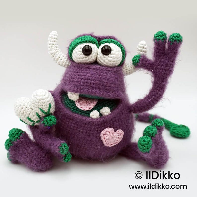 Puede incluir: Un juguete de monstruo tejido a ganchillo en color morado con detalles verdes, dos cuernos blancos y ojos grandes blancos con pupilas negras. Tiene una boca ancha con dientes, lengua rosa, un parche de coraz&oacute;n rosa y una cola larga. Texto: &copy; Ildikko www.ildikko.com