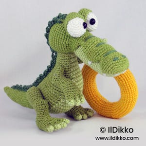 May include: A green crocheted crocodile toy with large white eyes and dark pupils, holding a bright yellow crocheted ring in its mouth. The crocodile has a textured back and tail. The text &copy; Ildikko www.ildikko.com is visible at the bottom.