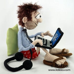 May include: A crocheted doll with brown spiky hair, wearing a light blue shirt, a dark tie, and red patterned shorts. The doll sits while using a black laptop with a blue screen and white text, wearing brown sandals, with black headphones nearby.