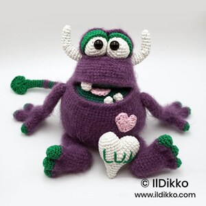Puede incluir: Un monstruo de ganchillo morado con dos cuernos blancos, grandes ojos blancos con pupilas negras y detalles verdes en pies, manos y cola. Presenta un parche de coraz&oacute;n rosa, un coraz&oacute;n blanco con el texto verde LUV y una boca ancha con dientes.