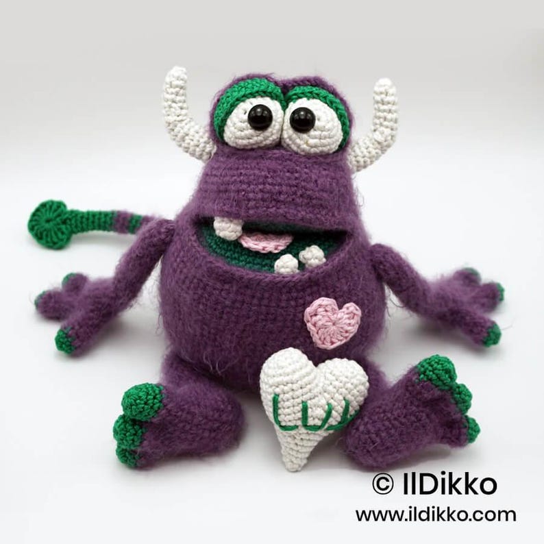 Puede incluir: Un monstruo de ganchillo morado con dos cuernos blancos, grandes ojos blancos con pupilas negras y detalles verdes en pies, manos y cola. Presenta un parche de coraz&oacute;n rosa, un coraz&oacute;n blanco con el texto verde LUV y una boca ancha con dientes.