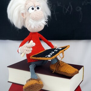 Puede incluir: Mu&ntilde;eco de ganchillo de Albert Einstein, con pelo y barba blancos, con un jersey rojo, pantalones grises y zapatos marrones. Sostiene una tiza y una peque&ntilde;a pizarra. El mu&ntilde;eco est&aacute; sentado sobre dos libros.