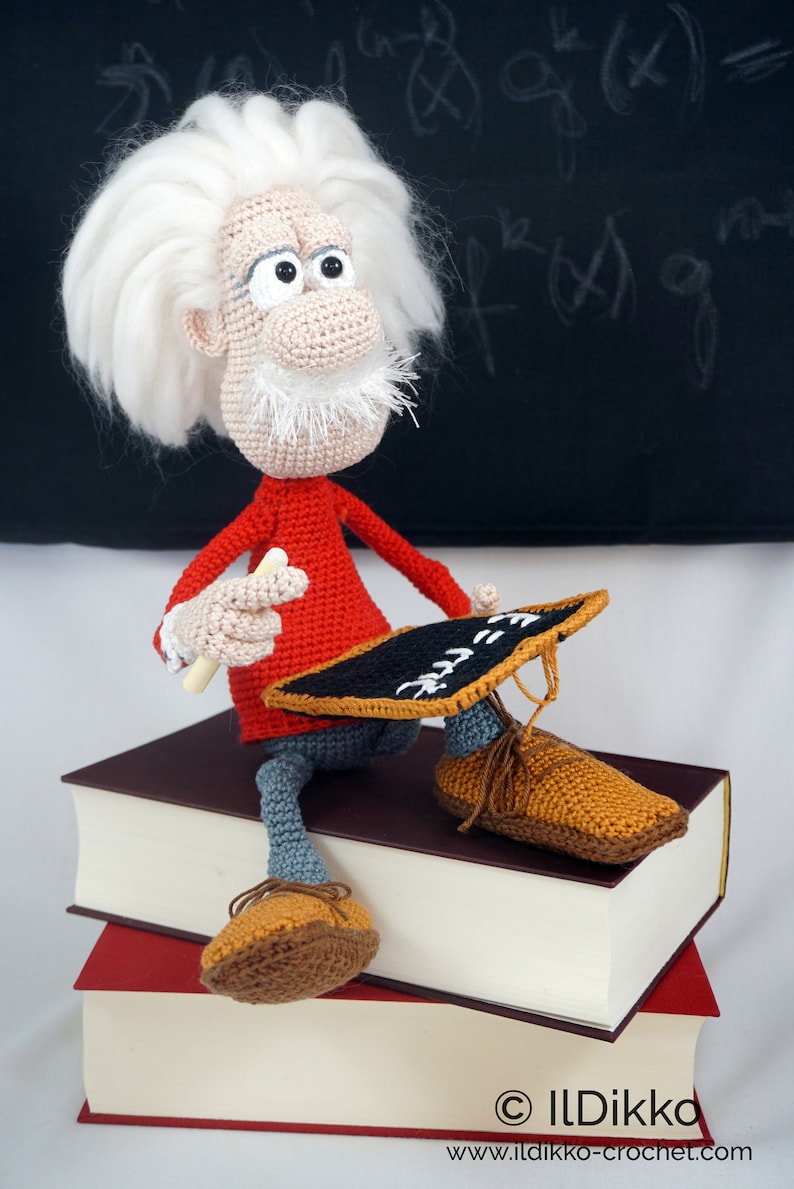 Puede incluir: Mu&ntilde;eco de ganchillo de Albert Einstein, con pelo y barba blancos, con un jersey rojo, pantalones grises y zapatos marrones. Sostiene una tiza y una peque&ntilde;a pizarra. El mu&ntilde;eco est&aacute; sentado sobre dos libros.