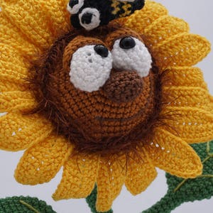 May include: A crocheted sunflower with a brown face, white eyes, and a black bee on top. The sunflower has yellow petals and a green stem with leaves.
