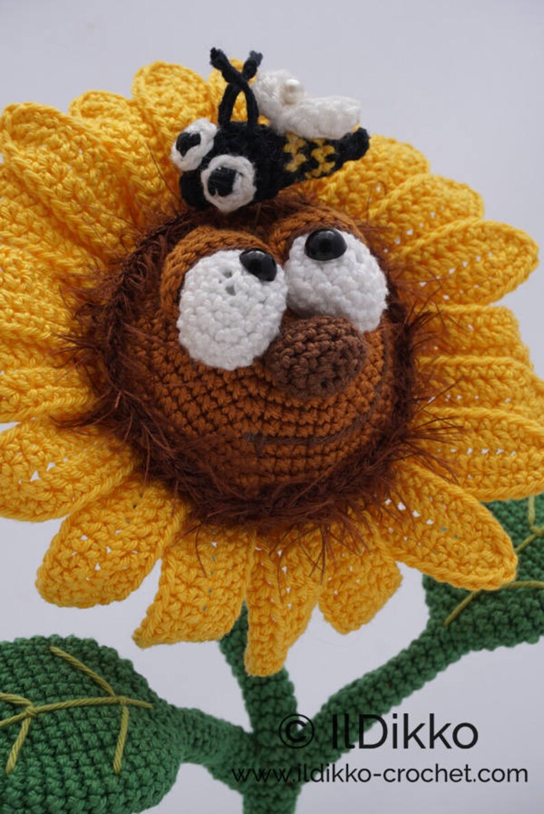 May include: A crocheted sunflower with a brown face, white eyes, and a black bee on top. The sunflower has yellow petals and a green stem with leaves.