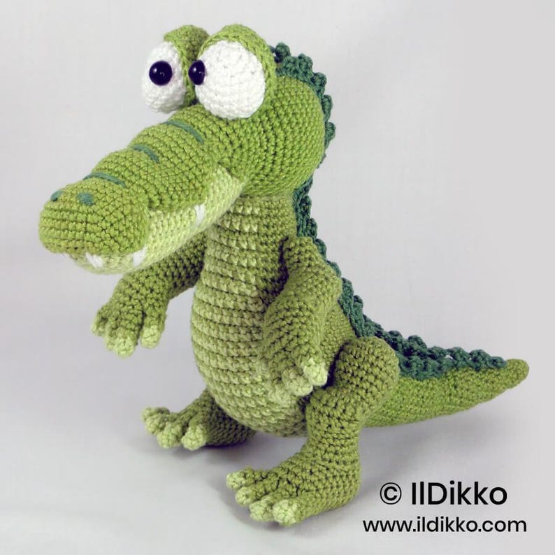 May include: A green crochet crocodile toy with large white eyes and black pupils. The creature features a textured back with dark green scales, a light green belly, and short limbs. The text "&copy; IlDikko www.ildikko.com" is visible in the corner.