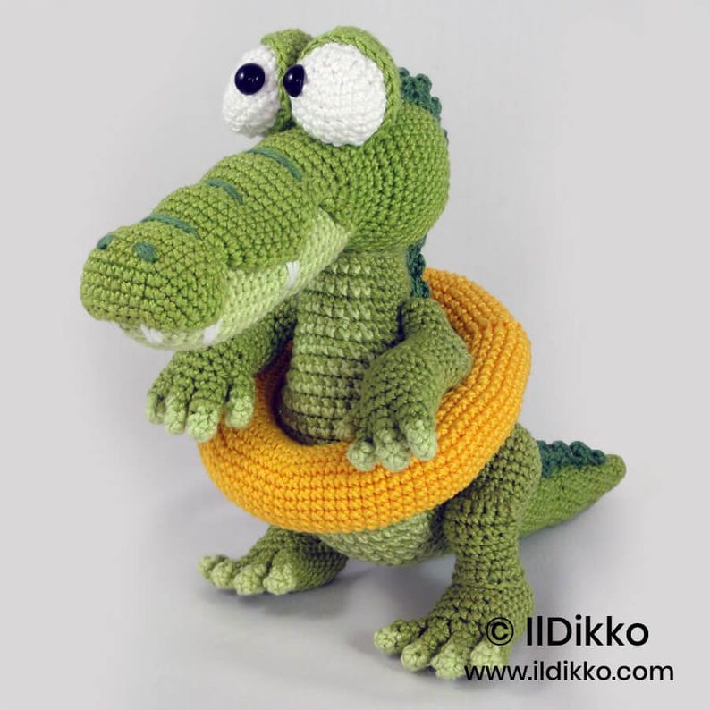 May include: A green crocheted crocodile stuffed animal with large white eyes and black pupils. The crocodile is wearing a bright yellow crocheted inner tube around its midsection. The text "Ildikko www.ildikko.com" is printed in the bottom right corner.