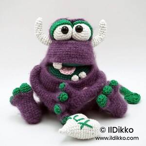 Puede incluir: Un mu&ntilde;eco monstruo de ganchillo morado con detalles verdes, ojos blancos grandes con pupilas negras, dos cuernos blancos y una boca ancha que muestra los dientes. Sostiene un diente de ganchillo blanco con costuras verdes. Texto: &copy; IlDikko www.ildikko.com