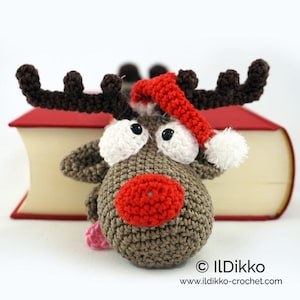 Puede incluir: Un juguete de reno tejido a ganchillo con cabeza marrón, una nariz roja grande y ojos blancos. Lleva un gorro de Papá Noel rojo y blanco y tiene astas de color marrón oscuro. El texto "ILDIKKO www.ildikko-crochet.com" aparece abajo a la derecha.