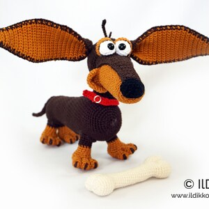 May include: A handmade crochet dachshund dog with large, floppy ears. The dog is brown and tan with a red collar and large, googly eyes. A cream-colored bone lies in front of the dog. The dog is a whimsical, handcrafted item.