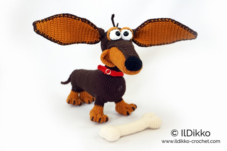 May include: A handmade crochet dachshund dog with large, floppy ears. The dog is brown and tan with a red collar and large, googly eyes. A cream-colored bone lies in front of the dog. The dog is a whimsical, handcrafted item.
