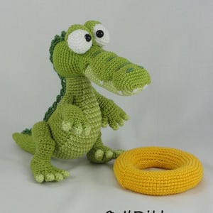 May include: A green crocheted crocodile toy with large white eyes and a dark green spine, standing next to a yellow crocheted ring. The text "&copy; ILDikko" and "www.ildikko-crochet.com" is printed in black at the bottom of the image.