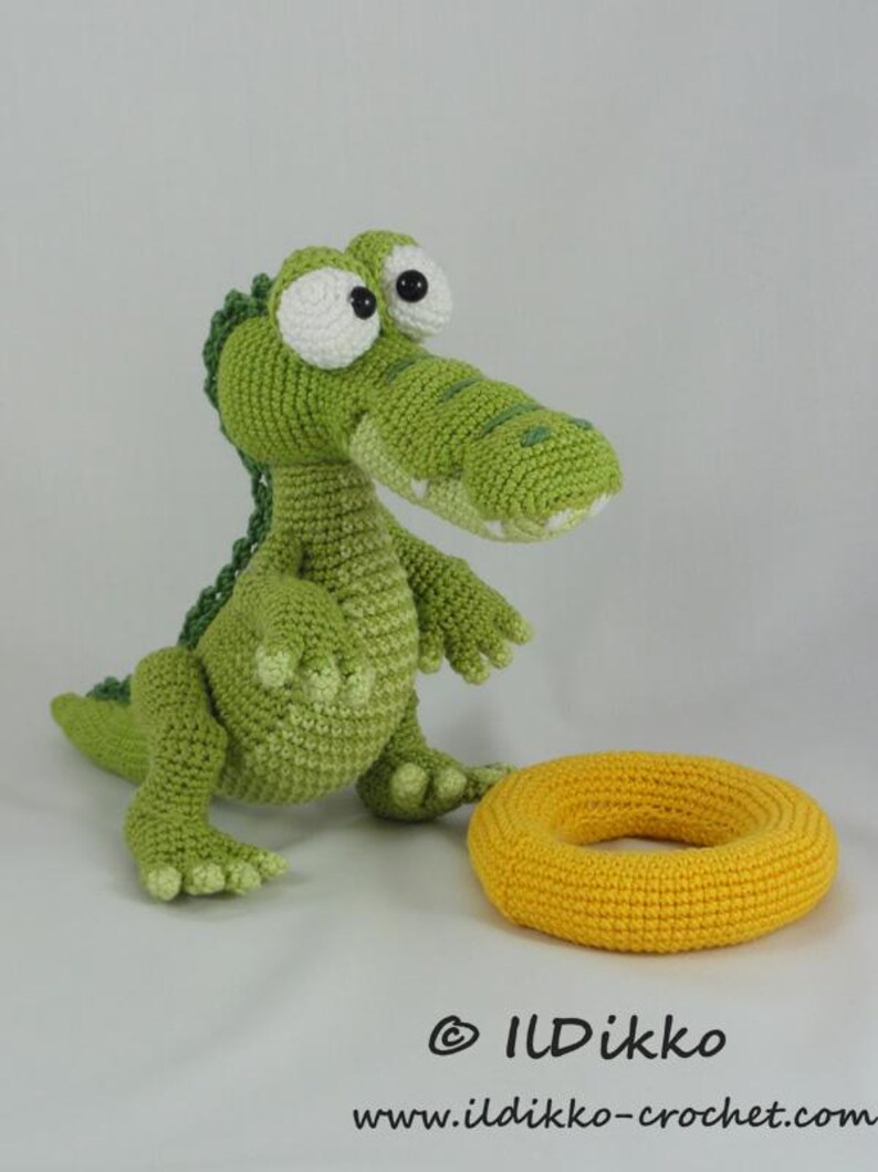 May include: A green crocheted crocodile toy with large white eyes and a dark green spine, standing next to a yellow crocheted ring. The text "&copy; ILDikko" and "www.ildikko-crochet.com" is printed in black at the bottom of the image.