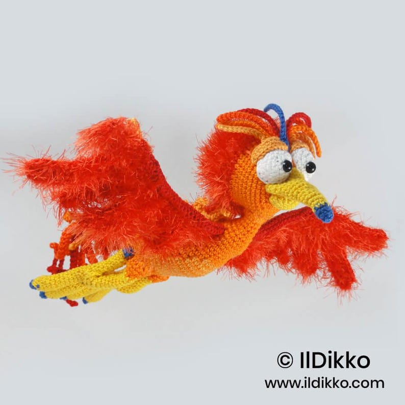 May include: A crocheted phoenix toy featuring vibrant orange and red yarn with a fuzzy texture on the wings. The bird has a yellow beak with a blue tip, large white eyes with black pupils, and a blue crest. Text reads: &copy; IlDikko www.ildikko.com.