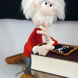 Puede incluir: Mu&ntilde;eco de Einstein tejido a ganchillo con pelo blanco, chaqueta roja y zapatos marrones. El mu&ntilde;eco sostiene un trozo de tiza y posa junto a una peque&ntilde;a pizarra. El mu&ntilde;eco est&aacute; sentado sobre una pila de libros.
