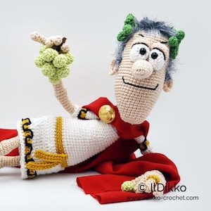 Op de afbeelding: Een gehaakte Caesar-pop, met een rode cape, witte tuniek met gouden rand en een lauwerkrans. De pop houdt een tros groene druiven vast. De pop heeft een glimlachende uitdrukking en ligt. De pop heeft een gouden knoop op de cape.