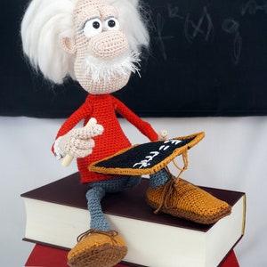 May include: A crocheted doll with white hair and a mustache, wearing a red long-sleeve shirt and gray pants, sits on two stacked books. The doll holds a small black chalkboard with white text and a piece of white chalk in its crocheted hands.