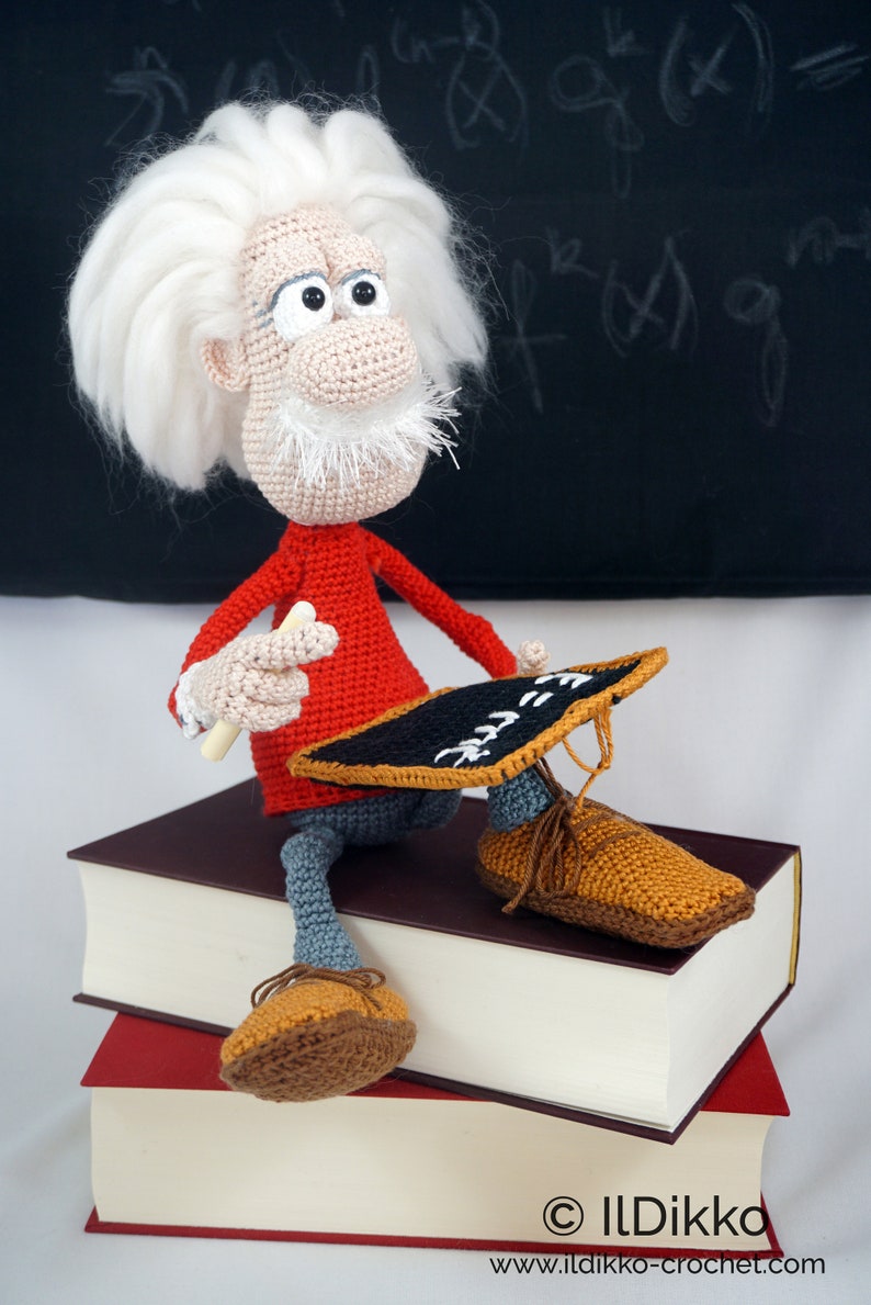 May include: A crocheted doll with white hair and a mustache, wearing a red long-sleeve shirt and gray pants, sits on two stacked books. The doll holds a small black chalkboard with white text and a piece of white chalk in its crocheted hands.