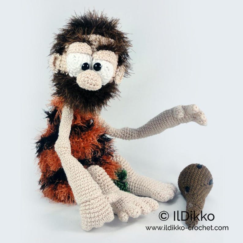 May include: A crocheted caveman doll with a shaggy dark brown beard and hair, wearing a textured orange and black tunic. The doll has large white eyes with black pupils, light beige skin, and is positioned next to a small brown club.