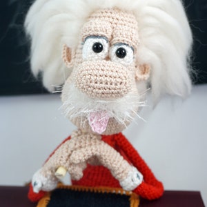May include: A crocheted doll featuring a beige face with a large nose, black eyes, and a pink tongue sticking out. The doll has wild white hair, a white mustache, and wears a red garment. It holds a small black chalkboard with E=mc&sup2; written in white.
