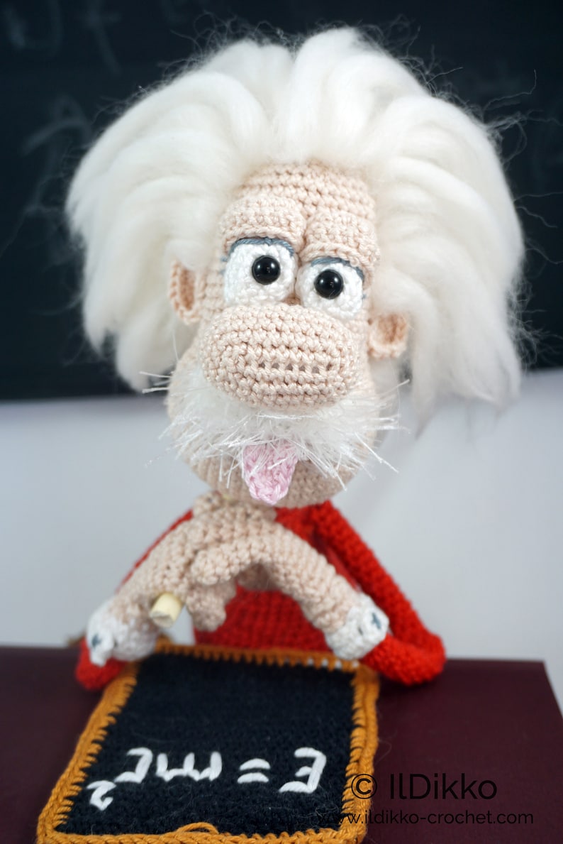 May include: A crocheted doll featuring a beige face with a large nose, black eyes, and a pink tongue sticking out. The doll has wild white hair, a white mustache, and wears a red garment. It holds a small black chalkboard with E=mc&sup2; written in white.