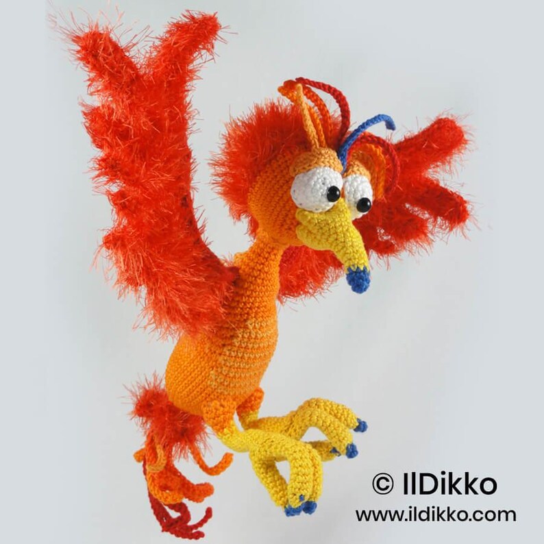 May include: A crocheted phoenix toy with bright orange and red fuzzy wings, a yellow body, and large white eyes with black pupils. It features a yellow beak with a blue tip, blue-tipped claws, and red and blue head crests. Text reads: &copy; IlDikko www.ildikko.com