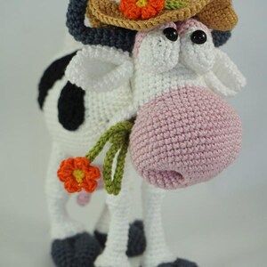May include: A crocheted cow wearing a brown straw hat with an orange flower. The cow has black and white spots and a pink nose. The cow is holding a green stem with an orange flower.