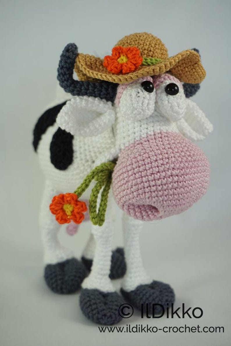 May include: A crocheted cow wearing a brown straw hat with an orange flower. The cow has black and white spots and a pink nose. The cow is holding a green stem with an orange flower.
