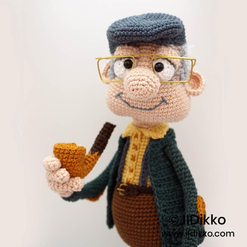 May include: A handmade crochet doll of an elderly man, wearing a blue cap, gold-rimmed glasses, and a dark green jacket. The doll holds a brown pipe and has a smiling expression. The doll is wearing a yellow shirt and brown pants.