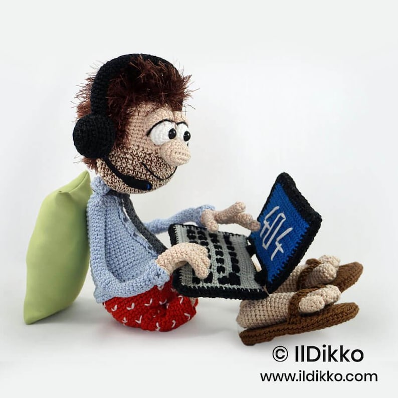 May include: A crocheted doll with brown hair, wearing a light blue shirt, red patterned shorts, and brown sandals. The doll has black headphones with a microphone, sits against a light green pillow, and holds a black laptop displaying 404.