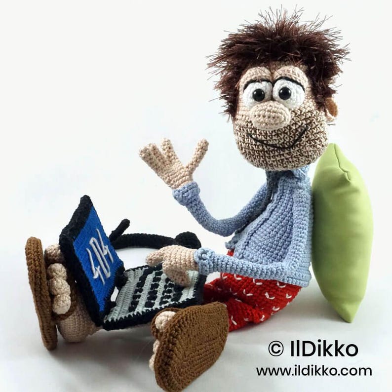May include: A crocheted doll with brown hair, a light blue shirt, and red patterned pants, sitting and using a black laptop with a blue screen displaying the text 404. The doll wears brown sandals and leans against a light green pillow.