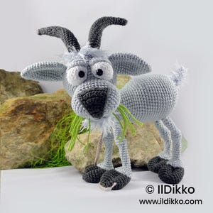 Può includere: Un giocattolo a forma di capra realizzato all'uncinetto in grigio chiaro con corna e zoccoli grigio scuro. Presenta grandi occhi bianchi con pupille nere, un naso grigio scuro e un ciuffo di peli bianchi sulla testa. Masticando fili di lana verde.