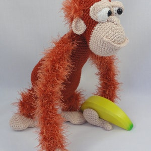 May include: A crocheted orangutan toy with a long, reddish-orange fur, holding a yellow banana. The toy has white eyes and a pink nose.