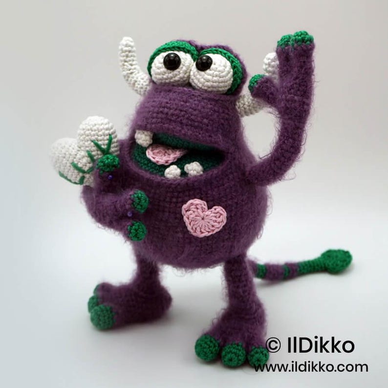 Puede incluir: Un monstruo de ganchillo morado con detalles verdes, cuernos blancos y ojos grandes. Sostiene un coraz&oacute;n blanco, tiene un coraz&oacute;n rosa en el pecho y una cola larga. El texto "&copy; IlDikko www.ildikko.com" aparece en la esquina inferior derecha.