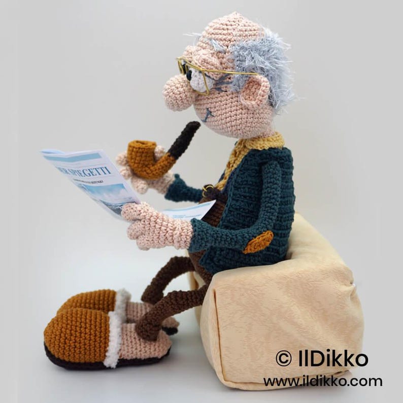 May include: Crocheted figure of an elderly man reading a newspaper and smoking a pipe. The figure wears glasses, a blue jacket, and brown slippers. The newspaper reads "R.EGETTI". The figure is seated on a beige cushion.