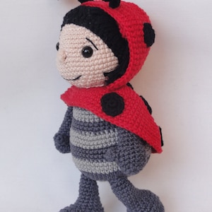 Pode incluir: Uma boneca de crochê em forma de joaninha com detalhes em preto e vermelho. A boneca tem um corpo listrado cinza e branco e olhos pretos.