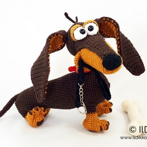 May include: A handmade crochet dachshund dog in brown and tan with large googly eyes, a red collar, and a black leash. A white bone is next to the dog. The dog is a whimsical, handcrafted item.
