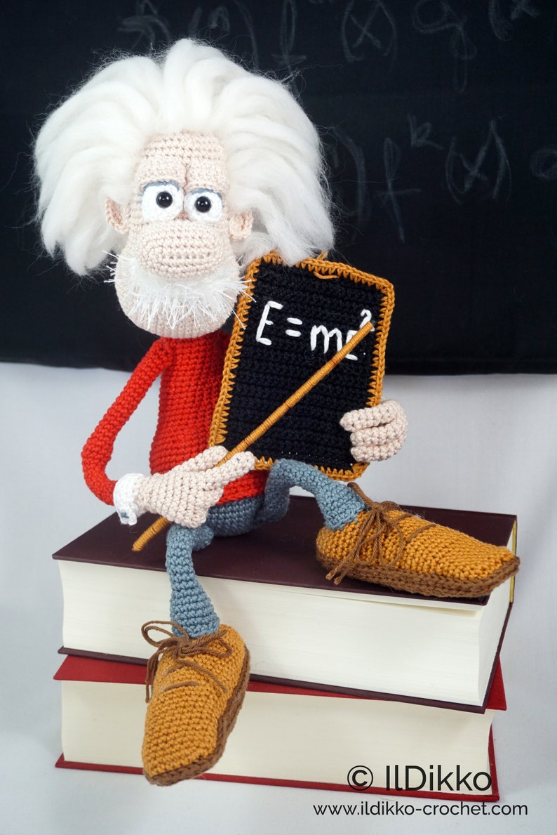 May include: A crocheted doll of a scientist with wild white hair, a red long-sleeved shirt, and gray trousers. The doll sits on two stacked books, holding a small black chalkboard with the white text E=mc&sup2; and a thin wooden pointer stick.